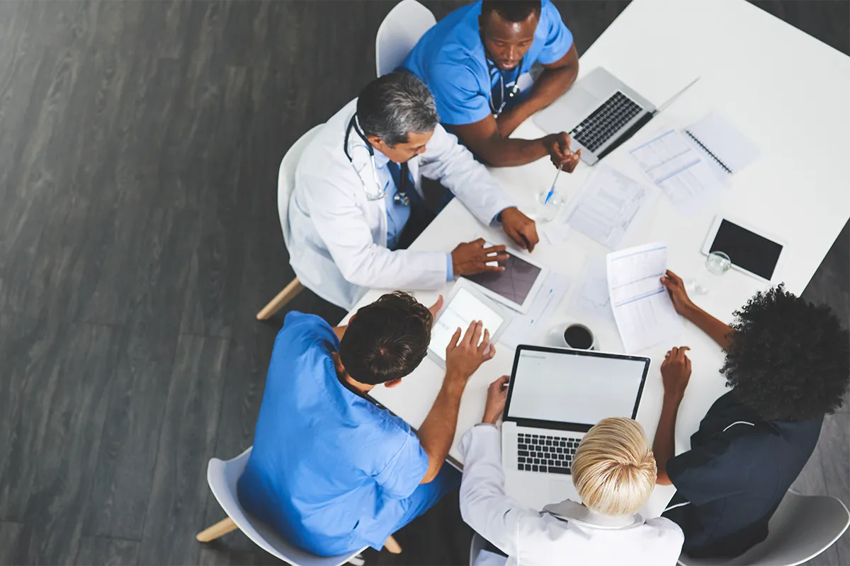 Équipe de professionnels de santé réunie autour d’une table avec ordinateurs, tablettes et documents.